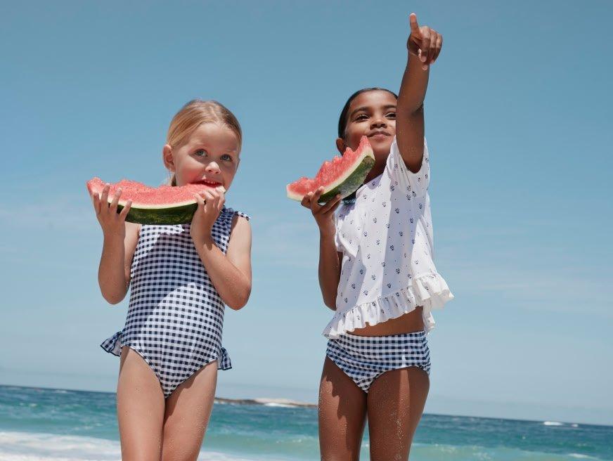 two little girls standing on the beach eating watermelon
