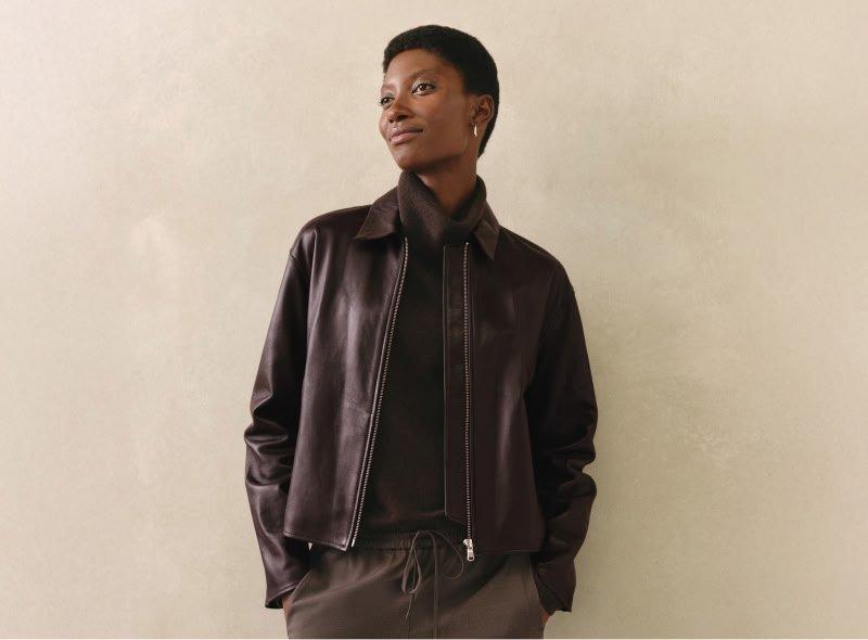 a woman in a brown leather jacket and pants leaning against a wall