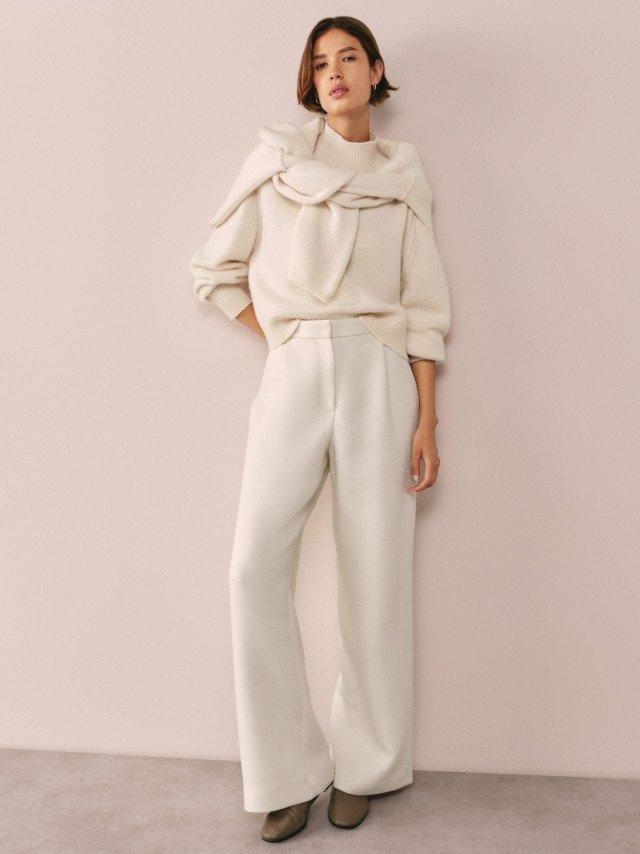 a woman in white pants and a white top posing against a wall