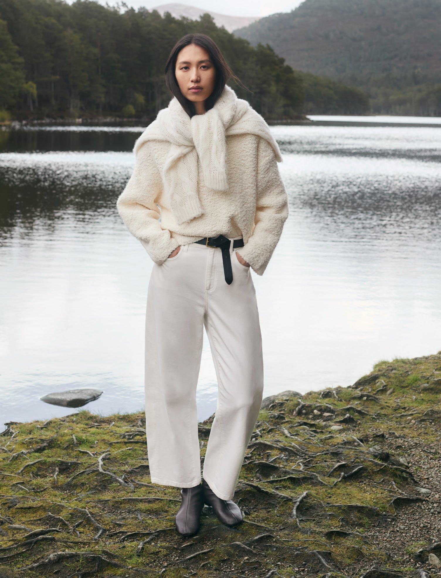a woman in white pants and a white sweater standing by a lake