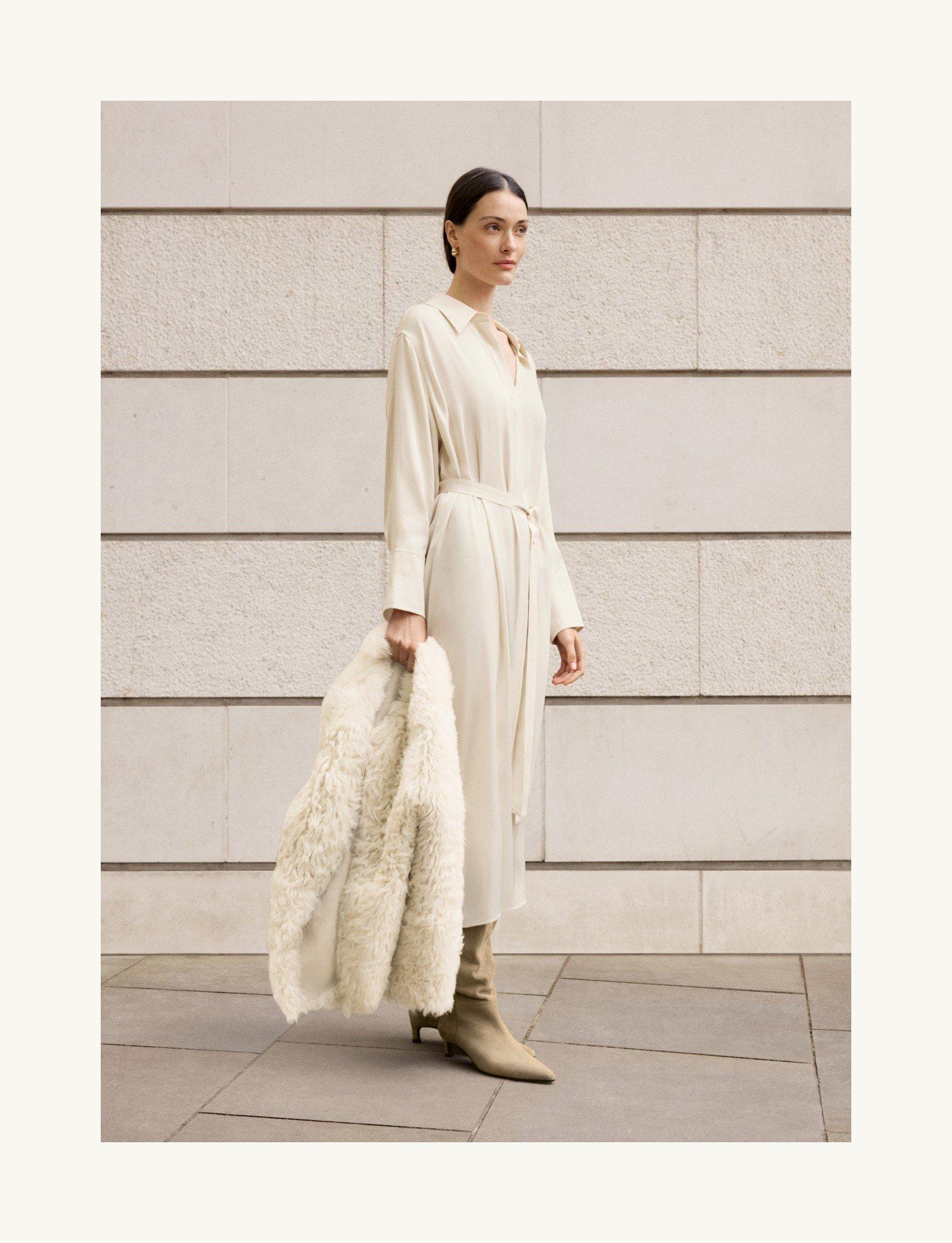 a woman in a white dress and fur coat standing on a sidewalk