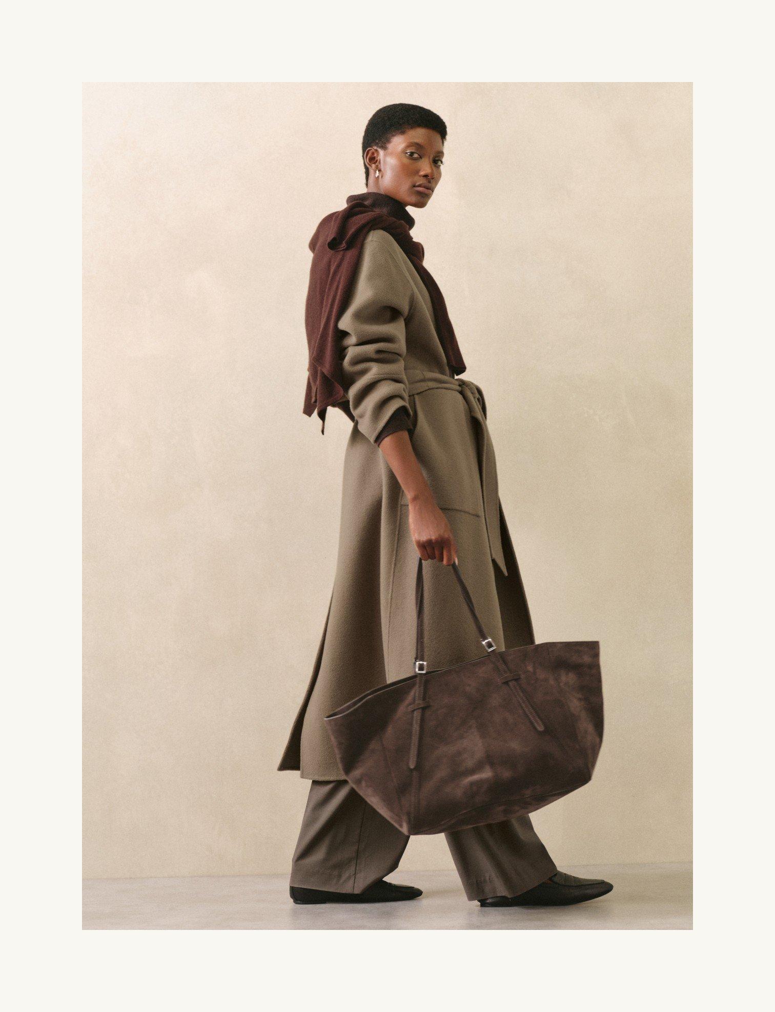 a woman in a trench coat and brown boots holding a large brown bag