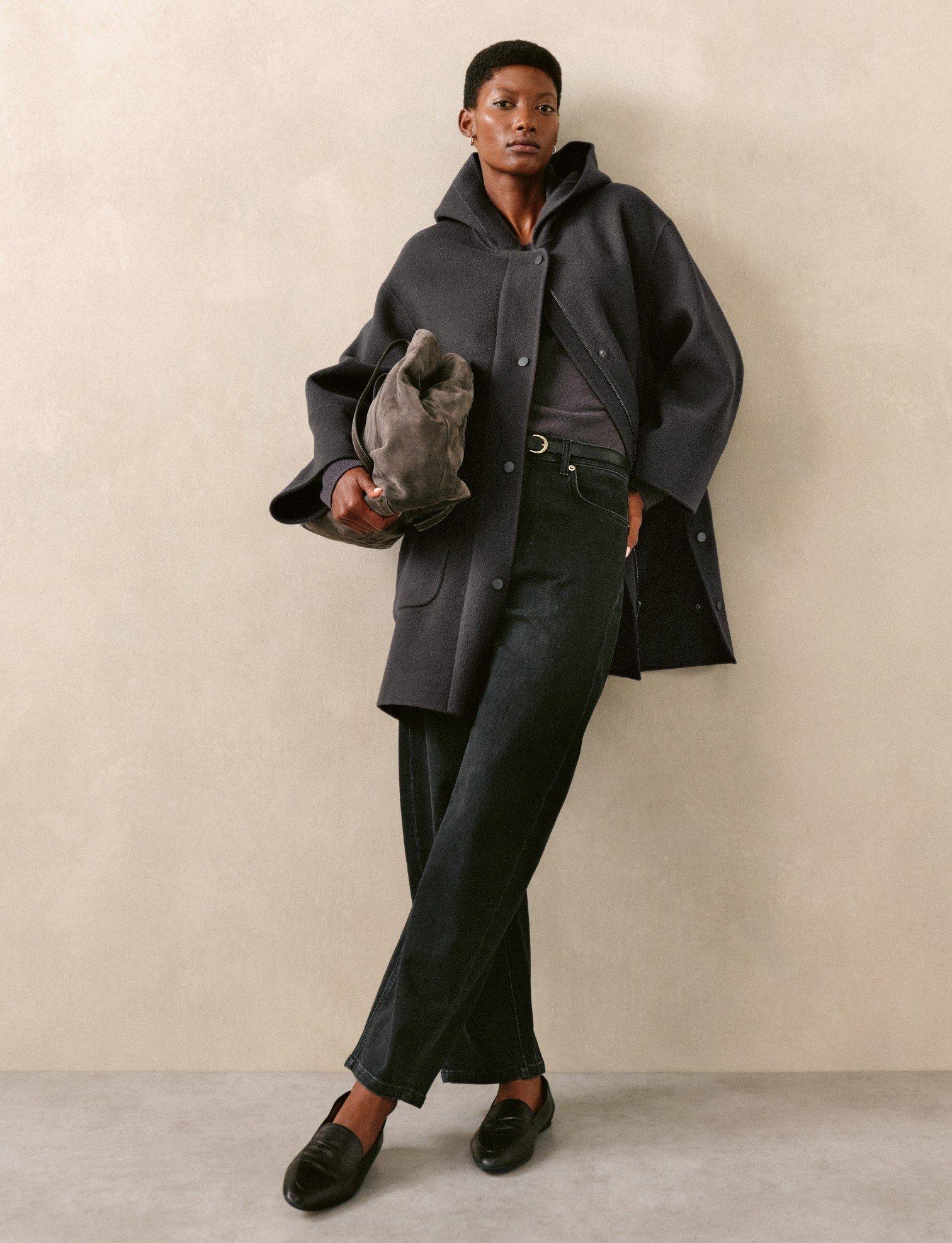a woman in a black coat and pants leaning against a wall