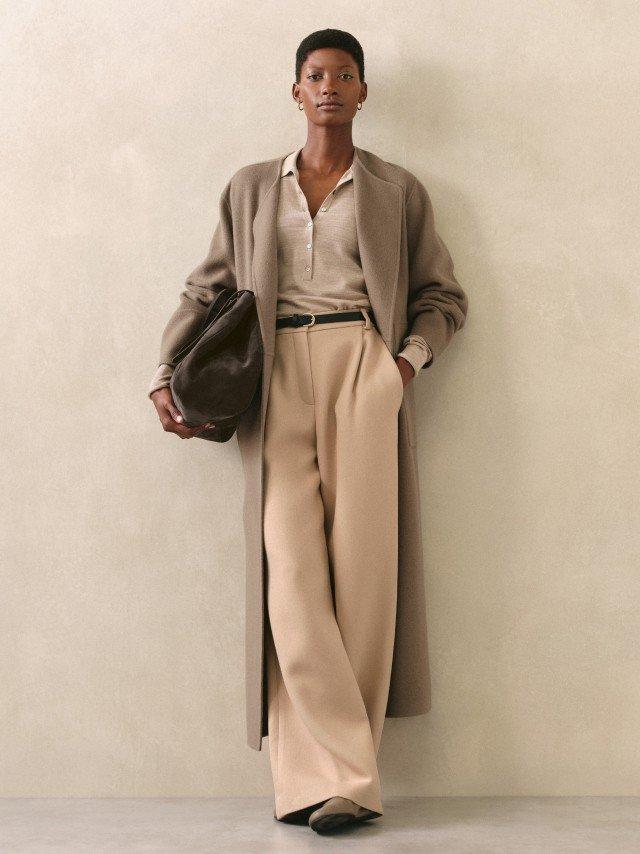a woman in a beige coat and pants standing against a wall