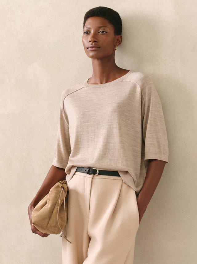 a woman in beige pants and a tan top leaning against a wall