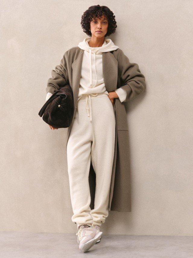 a woman in a white coat and beige pants leaning against a wall