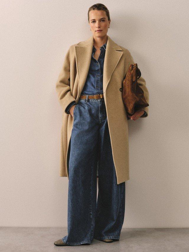 a woman in a beige coat and jeans standing against a wall