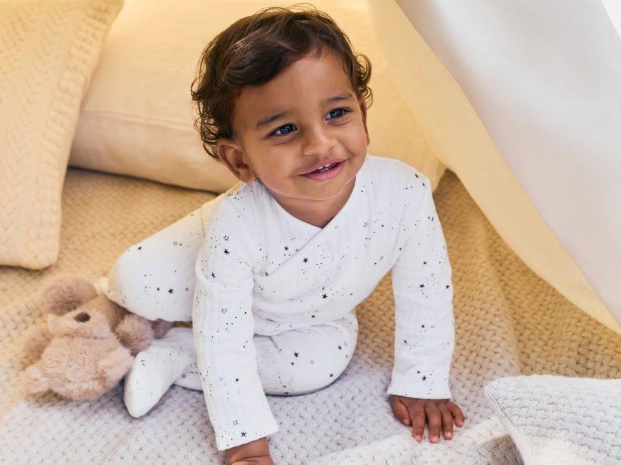 a baby in white pajamas sitting on a bed with a teddy bear